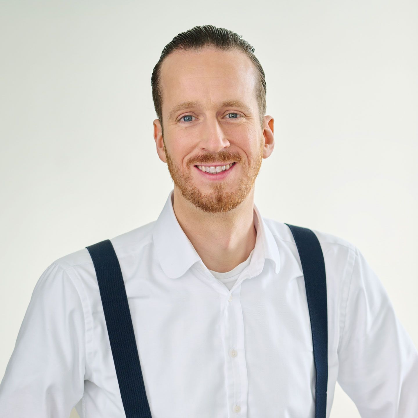 Sebastian Pollmer, muškarac stoji u bijeloj košulji s plavim tregerima u svijetloj prostoriji. 