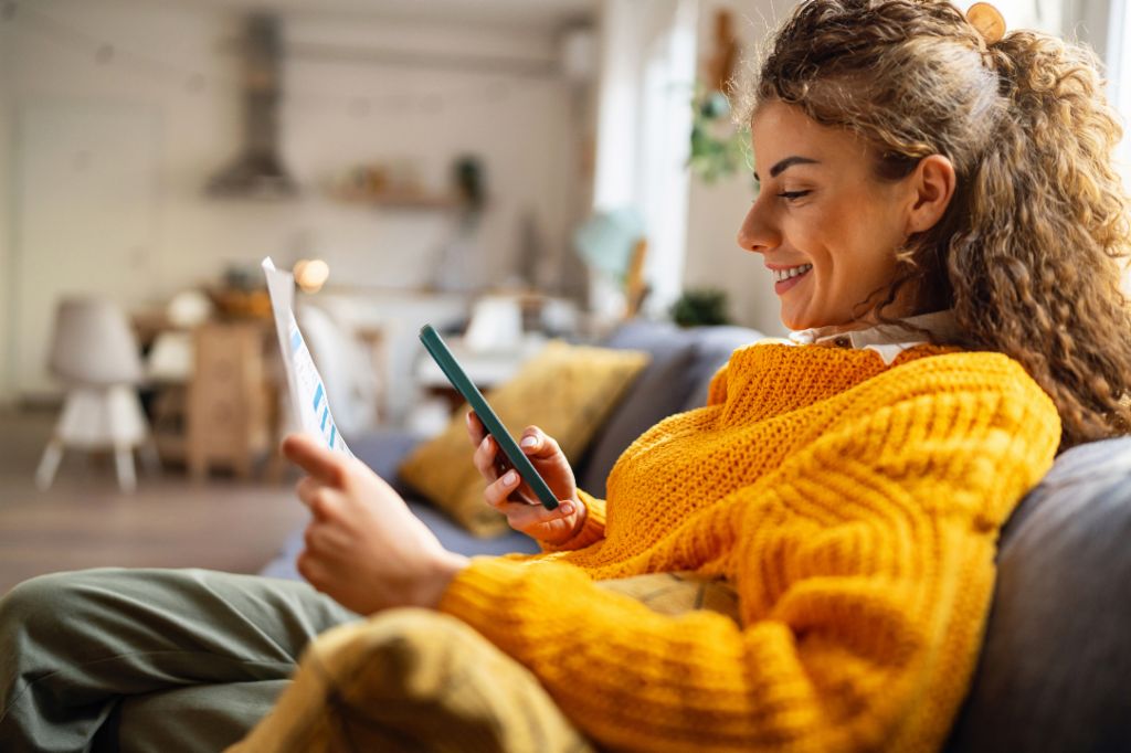 Nasmijana mlada žena s kovrdžavom kosom, u udobnom žutom džemperu, sjedi na kauču u svojoj dnevnoj sobi i gleda u svoj pametni telefon dok drži papirni dokument.