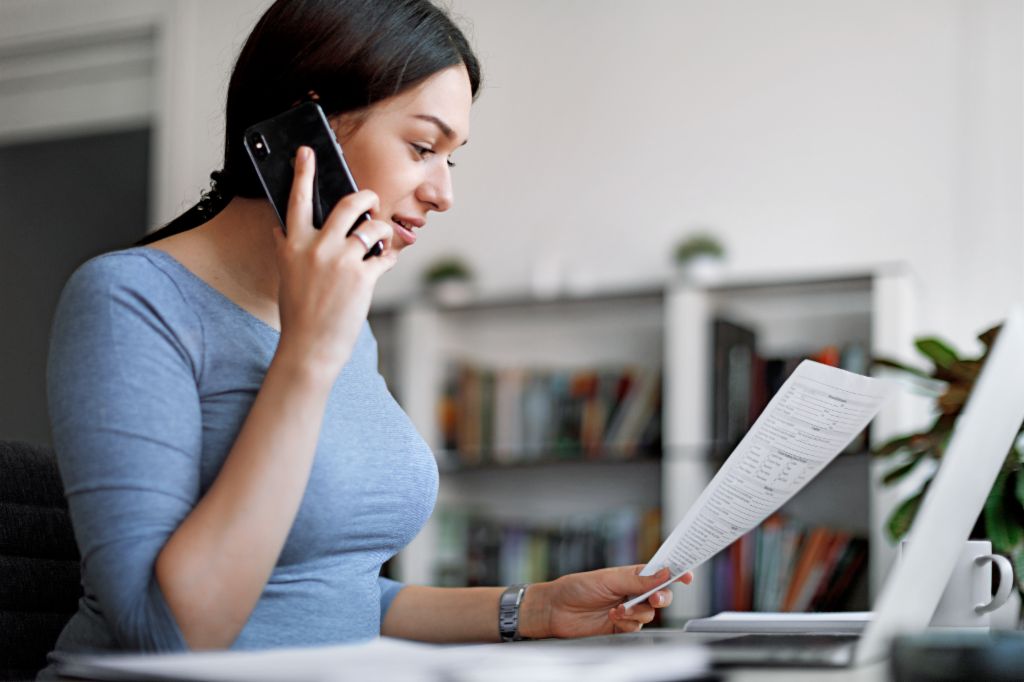 Žena u plavoj majici na mobilnom telefonu dok drži račun u rukama.
