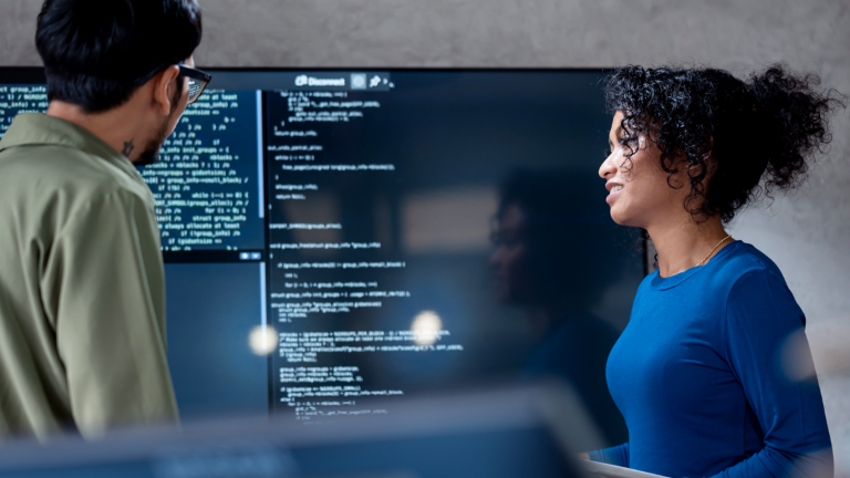 AI u naplati dugova: Na ovoj slici, dva stručnjaka zajednički analiziraju programski kod za automatizovane procese na velikom ekranu. Scena ilustruje praktičnu primenu veštačke inteligencije (AI) i robotizovane automatizacije procesa (RPA) u upravljanju dugovima – primer digitalne transformacije u upravljanju potraživanjima.
