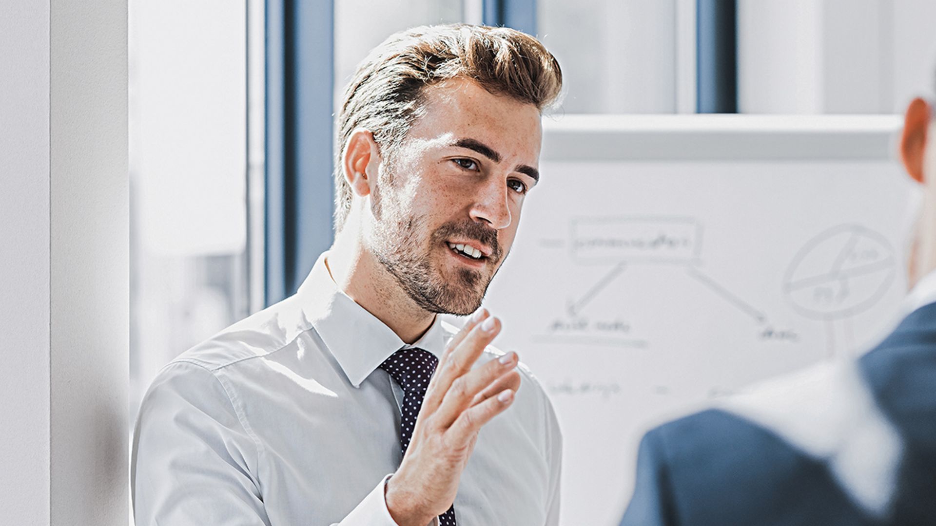 Vidi se muškarac u poslovnoj odjeći kako nešto objašnjava s prijateljskim i angažiranim izrazom lica. Ispred njega stoji druga osoba koja ga očito sluša. U pozadini se vidi bijela tabla sa skicom.