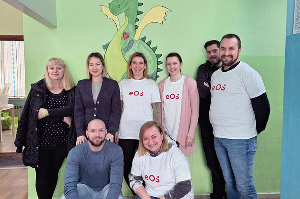 Osam ljudi stoji ispred zida na kojem je naslikan zeleni zmaj. Ljudi su raspoređeni u dva reda: dvoje čuče naprijed, a šestoro stoji odzada. Četiri osobe nose bijele majice kratkih rukava sa crvenim logom kompanije EOS. Grupa se smiješi prema foto aparatu. Na podu, naprijed sa desne strane, vidimo nekoliko boca i kartonskih kutija sa pićem.