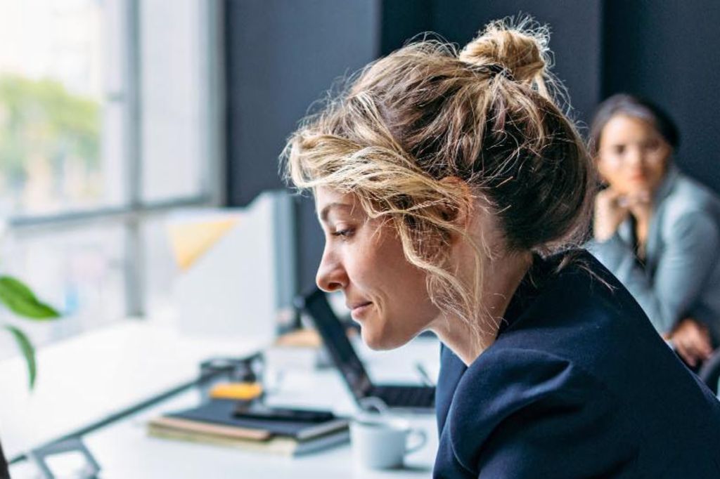 Slika prikazuje ženu koja sjedi za stolom u modernom uredu, obučena u tamni sako dok se fokusira na svoj laptop, s biljkom i nekim kancelarijskim priborom vidljivim na stolu, a u pozadini sjedi još jedna osoba.