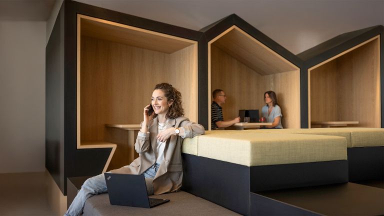 A smiling person sits in a modern office lounge, talking on the phone and working on a laptop. In the background, two other people are working.